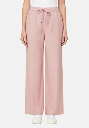 Donna che indossa pantaloni larghi di colore rosa chiaro con coulisse in vita e scarpe da ginnastica bianche, in piedi su uno sfondo neutro.