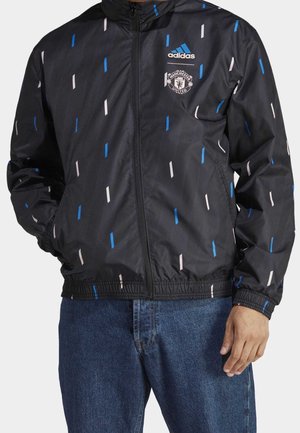 Black Adidas zip-up jacket with blue and white vertical dash patterns, Manchester United logo on chest, worn with blue jeans.