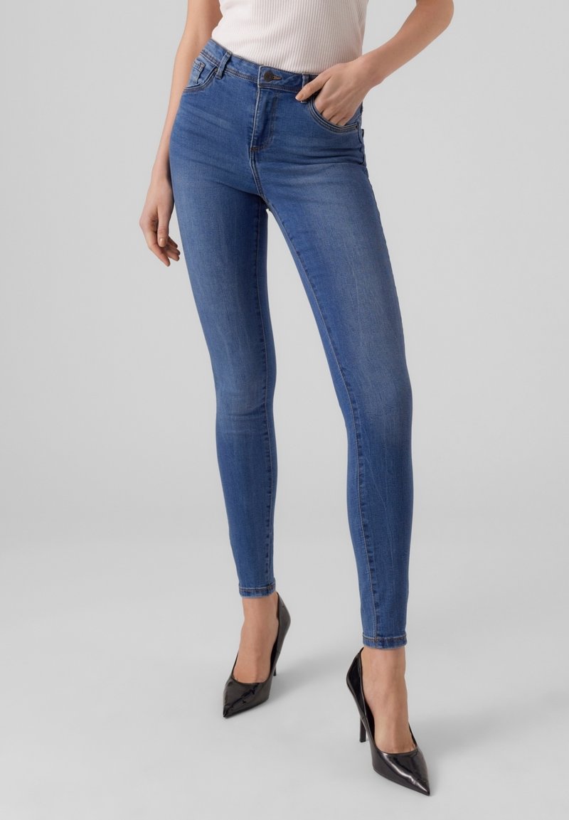 Femme portant un jean skinny bleu taille haute et des escarpins noirs en cuir verni à bout pointu, main dans la poche.