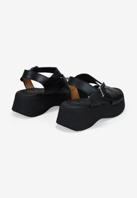 Sandalias de plataforma negras con tiras cruzadas, hechas de cuero. Cuentan con un cierre de hebilla y una suela gruesa y texturizada.