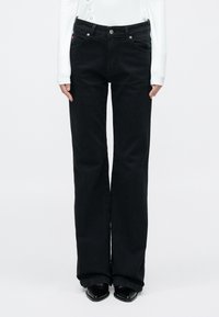 Fekete farmere bővülő szárú nadrág magas derékkal, gombos záródással és oldalzsebekkel. A modell fehér hosszú ujjú felsőt és fekete cipőt visel.