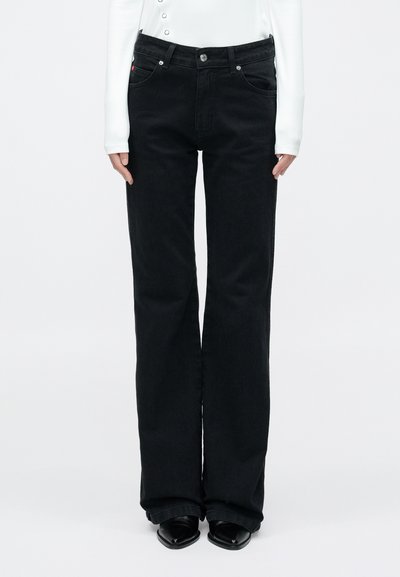 Pantalon évasé en denim noir avec taille haute, fermeture à boutons et poches latérales. Le mannequin porte un haut blanc à manches longues et des chaussures noires.