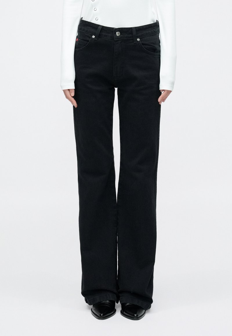 Fekete farmere bővülő szárú nadrág magas derékkal, gombos záródással és oldalzsebekkel. A modell fehér hosszú ujjú felsőt és fekete cipőt visel.