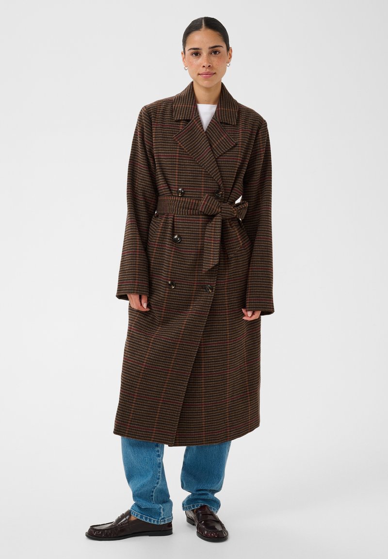 Femme portant un manteau marron à carreaux croisé avec ceinture, jean bleu et mocassins brun foncé, debout devant un fond uni.