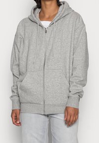 Sweat à capuche gris zippé en tissu doux avec capuche, poches avant et poignets côtelés, doté d'une coupe décontractée et de longs cordons de serrage.