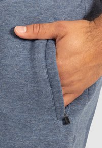 La tela azul de mezcla de algodón cuenta con un bolsillo con cremallera. Una mano descansa dentro del bolsillo, mostrando la suavidad del tejido.