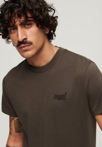 Camiseta de algodón marrón con mangas cortas, escote redondo y un pequeño logo bordado en azul marino en el lado izquierdo del pecho.