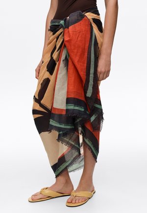 Personne portant un sarong multicolore à motif abstrait noué à la taille avec des sandales beiges sur fond blanc.