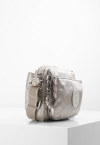Small bag in metallic beige material with a front logo patch, dual zipper closures, and a canvas strap with chain accents.