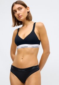 Schwarzer Bralette mit V-Ausschnitt und breitem weißen Bündchen mit dem Logo 'Calvin Klein'; kombiniert mit passenden schwarzen Unterhosen. Glatte Textur.