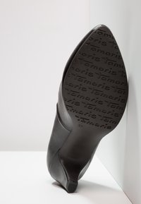Chaussure noire à talons hauts avec tige en cuir lisse et semelle en caoutchouc texturée ornée d’un logo en relief. Bout pointu et talon fin.