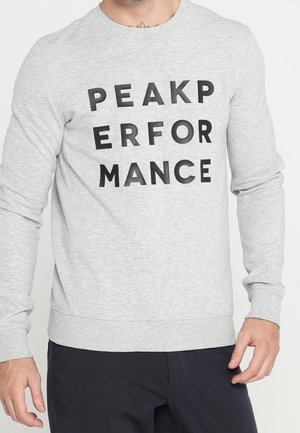 Mężczyzna ubrany w jasnoszarą bluzę z czarnymi grubymi literami tworzącymi napis „PEAK PERFORMANCE” w trzech liniach z przodu.