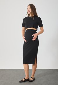 Μαύρο ριμπ μεταφορικό ρούχο δύο τεμαχίων που περιλαμβάνει ένα κοντό τοπ και μια φούστα μέχρι το γόνατο με πλαϊνό σκίσιμο. Το μοντέλο φορά μαύρες μπαλαρίνες.