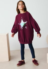 Un bambino sta in piedi con sicurezza, indossando una felpa oversized color bordeaux con un design a stella, abbinata a leggings blu navy e sneaker bordeaux.