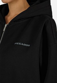 Κοντινό πλάνο ενός ατόμου που φοράει ένα μαύρο φούτερ με φερμουάρ και κουκούλα, με τη λέξη "PEGADOR" τυπωμένη σε γκρι στο στήθος.