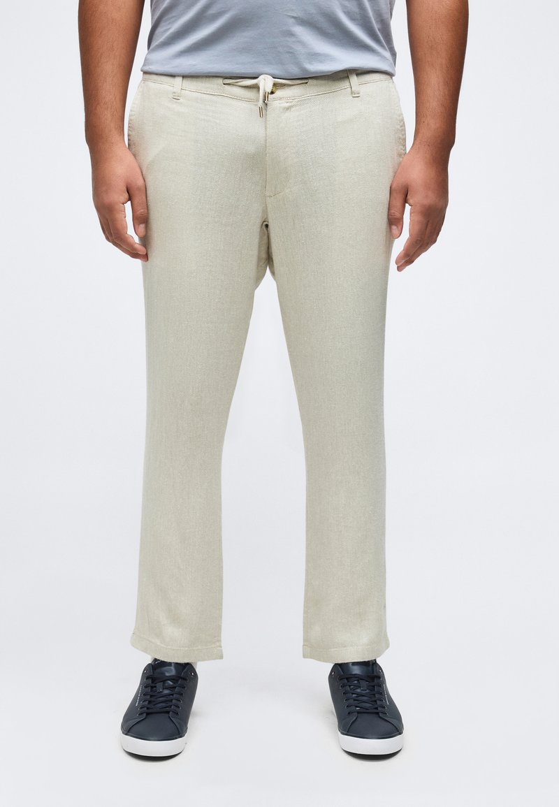 Férfi világos bézs, zsinóros nadrágban, szürke pólóban és fekete sportcipőben, fehér háttér előtt áll.