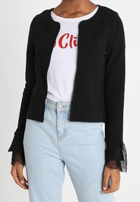 Zwarte geribbelde gebreide jas met een ronde hals, open voorkant en kantaccenten bij de mouwen, gedragen over een wit T-shirt en lichtblauwe jeans.