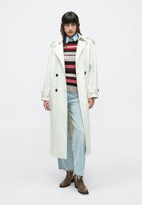 Hvid trenchcoat med store knapper, lagdelt over en flerfarvet striped sweater, kombineret med lyseblå slidte jeans og brune ankelstøvler.