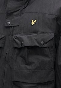 Černá lehká bunda s texturovaným povrchem, vybavená zipovým zapínáním, dvěma náprsními kapsami a zlatě vyšívaným logem na hrudi.