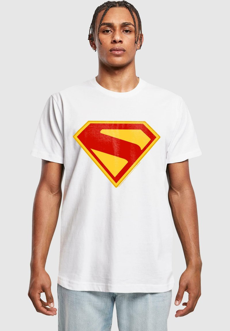 Jauns vīrietis ar pītām matiem, ģērbts baltā T-kreklā ar lielu sarkanu un dzeltenu stilizētu Supermena logo priekšpusē.
