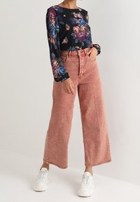 Blumenmuster-Bluse mit langen Ärmeln in Schwarz mit Schattierungen von Blau, Rosa und Grün. Kombiniert mit hochgeschnittenen, weit geschnittenen, ausgebleichten rosa Jeans und weißen Sneakers.