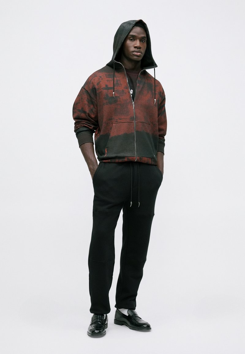 Mand iført en sort og rød tie-dye zip-up hoodie, sorte joggerbukser og sorte loafers står med hænderne i lommerne mod en hvid baggrund.