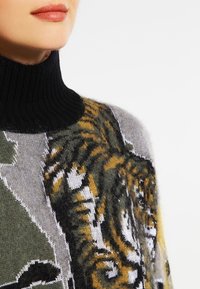 Gestrickter Pullover mit hohem, schwarzem, geripptem Kragen, mit einem Tigermotiv in Grau, Grün und Gelb auf einem weichen, strukturierten Körper.