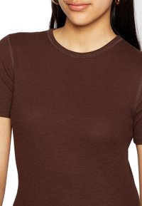 T-shirt castanho com nervuras, mangas curtas e decote redondo, apresentando uma textura suave e costuras contrastantes ao longo das bordas.