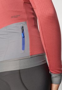 Langærmet top med en struktureret rød øverste sektion og en glat grå nederste panel. Har en lynlåslomme med en blå pull-tab.
