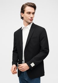 Νέος άνδρας με καλοχτενισμένα καστανά μαλλιά, φοράει μαύρο σακάκι πάνω από λευκό πουκάμισο και μπλε τζιν, προσαρμόζοντας το μανίκι του σακακιού του.