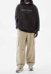 Felpa grigia scura con il testo "RHODE ISLAND", tasca a marsupio, abbinata a pantaloni cargo beige chiaro e sneakers grigie con motivi decorativi.