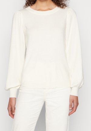 Femme portant un haut blanc uni à manches longues bouffantes et un pantalon blanc, debout devant un fond uni.