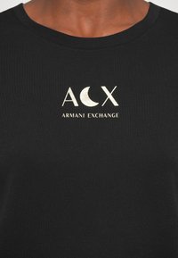 T-shirt de algodão preta com um logo impresso a ouro "ACX" e texto "Armani Exchange". Decote redondo com uma textura suave.
