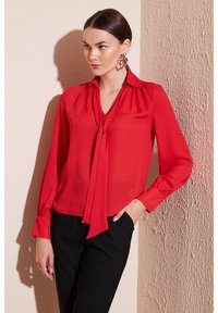 Red, long-sleeved blouse with a tie neckline, made of lightweight fabric, paired with black pants. Features gathered sleeves and a classic collar.