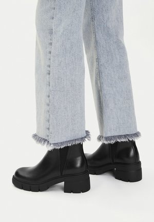 Jeans a vita alta con orlo sfrangiato in denim chiaro abbinati a stivaletti in pelle neri con suola spessa su sfondo bianco.