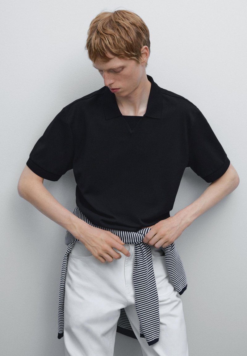 Černé polo tričko s designem ve tvaru V, s krátkými rukávy ohrnutými, v kombinaci s bílými džínami a svetrem pruhovaným v námořnickém stylu uvázaným v pase.