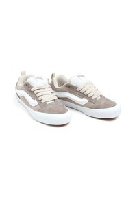 Zapatillas bajas de ante beige con acentos blancos, delantero con cordones y detalles texturizados. Cuentan con una suela de goma y un diseño que ofrece soporte.