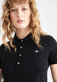 Černé polo triko vyrobené z texturované tkaniny s límečkem, knoflíkovou légou na tři knoflíky a malou vyšitou logem na levé části hrudi.