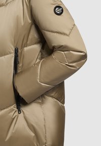 Padded winter jacket in beige with a smooth, shiny texture. Features diagonal stitching, a zippered pocket, and a logo patch on the sleeve.