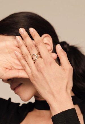 Personne aux cheveux foncés se couvre le visage avec les deux mains, montrant une bague en or au design incurvé sur le majeur gauche.