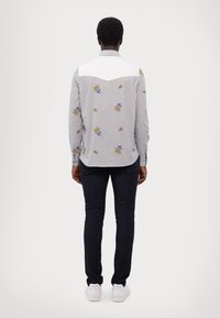 Camicia button-down grigia chiara con ricami floreali blu e gialli; spalle bianche e maniche lunghe. Abbinata a jeans scuri e sneakers bianche.