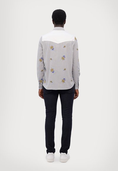 Camisa de botones en gris claro con bordados de flores azules y amarillas; con canesú blanco y mangas largas. Combinada con jeans oscuros y zapatillas blancas.