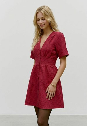Blonde woman smiling, wearing textured red short-sleeve dress with buttons and a bracelet, standing against plain light background.