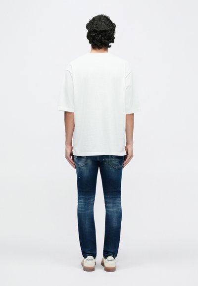 Maglietta bianca in cotone con maniche corte, abbinata a jeans in denim blu scuro con dettagli sbiaditi e tasche posteriori visibili. Scarpe da ginnastica visibili.