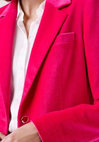 Blazer de terciopelo fucsia con solapas de muesca, bolsillo en el pecho y cierre de botón. Textura suave y ajuste entallado. Camisa blanca debajo.