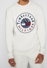 Felpa bianca con logo Timberland e Hilfiger ricamato circolare. Polsini e bordino a costine con una texture morbida.