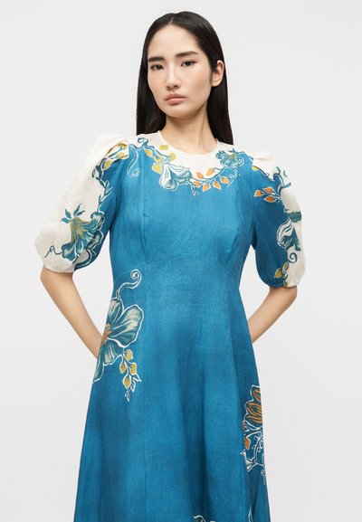 Γυναίκα με μακριά σκουρόχρωμα μαλλιά που φοράει μπλε φόρεμα με φουσκωτά λευκά μανίκια και μεγάλα λουλουδάτα σχέδια σε πράσινο, πορτοκαλί και κίτρινο.