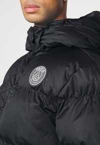 Homme portant une doudoune noire avec capuche et un logo du Paris Saint-Germain sur la poitrine.