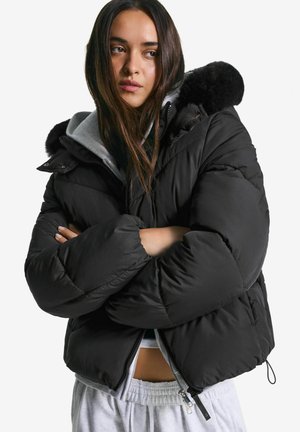 Jonge vrouw met lang donker haar, gekleed in een zwarte pufferjack met een bontgevoerde capuchon, een grijze hoodie en lichtgrijze joggingbroek, met gekruiste armen.