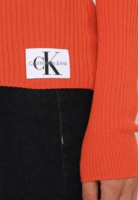 Ribbad orange tröjaärm med en vit etikett som har svart text "CALVIN KLEIN JEANS" och logotyp. Mörk denim syns nedanför.